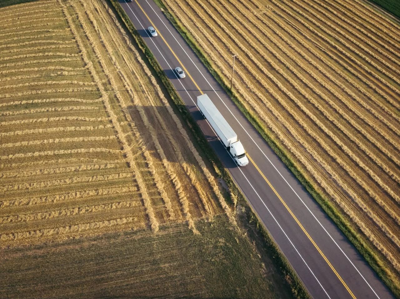 Tráiler blanco en una carretera entre sembradíos, visto desde arriba.